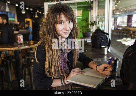 Portrait of smiling beautiful girl with dreadlocks and tattoos. Writing on a notebook with a pen in her hand. Selective focus with blurred background Stock Photo