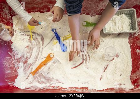 Close up shot from above of two pairs of kid hands playing with a sandy material on the colored table of the indoor play area. Experimenting Stock Photo