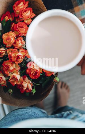 Woman holding white cup of coffee, latte cappucino, in background of bouquet orange roses flowers. Good morning concept. Spring romance. Breakfast Stock Photo