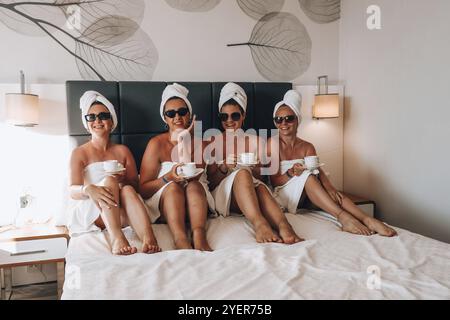 Four girls friends in white towels sitting on the bed having fun in hotel. Vacation Bloggers sunglasses drinking coffee together. Chat sweetly, tell s Stock Photo