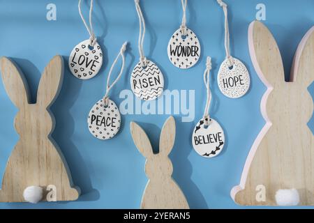 Group of wooden bunny ears and clay Easter eggs with words RISEN HOPE BELIEVE PEACE LOVE WONDER on blue background. Children air dry clay activity han Stock Photo