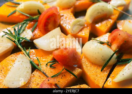Sliced pumpkin with onion, garlic and rosemary leaves on baking tray. Autumn food still life. Raw pumpkin slices with herbs and spices, close up. Stock Photo