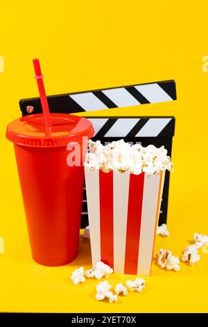 Vertical image of popcorn with red soda glass and white and black wooden clapperboard and scattered popcorn on a yellow background, entertainment and Stock Photo