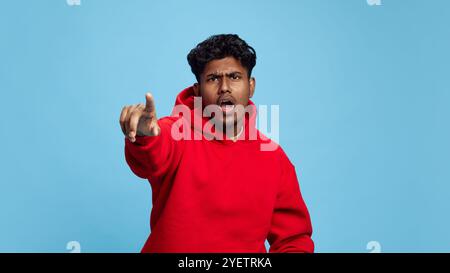 Emotional young Indian man wearing red hoodie, pointing forward with exclamation against blue studio background Stock Photo