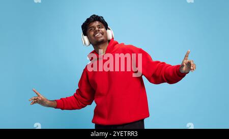 Happy, positive young Indian man in red hoodie, listening to music in headphones and dancing, having fun against blue studio background Stock Photo