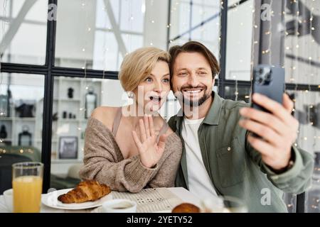 A loving couple enjoys breakfast together while taking a joyful selfie at home. Stock Photo