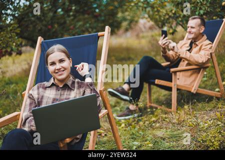 A cute woman in a shirt sits on a chair in the garden and works on a laptop. Female freelancer works remotely. Stock Photo