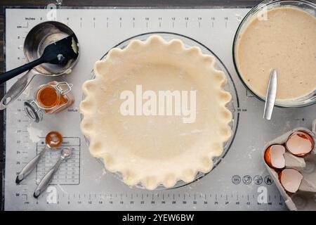 Unbaked Pie Crust with White Pumpkin Pie Filling Nearby: Overhead of fluted pie crust dough in glass pie pan with filling and equipment on the side Stock Photo