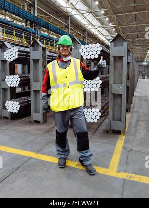 Worker with packs of rolled metal bars on the shelves holds thumbs up. Stock Photo