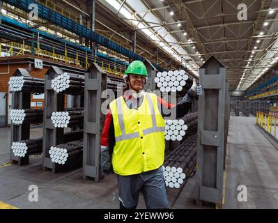 Worker with packs of rolled metal bars on the shelves holds thumbs up. Stock Photo