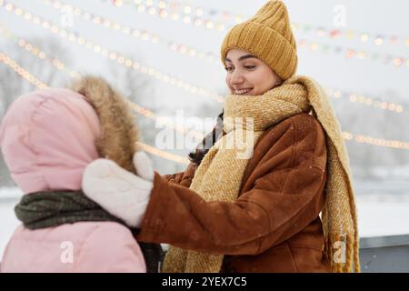 Side view portrait of caring young mother adjusting scarf on daughter in winter park and smiling happily with snow falling Stock Photo