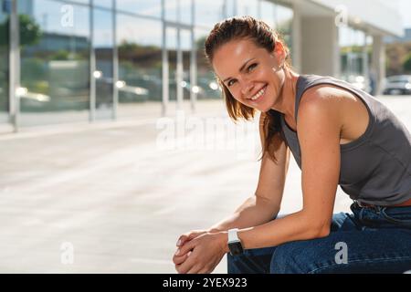 Portrait of happy young adult brunette woman sitting on a bench in an urban setting, smiling at the camera. Stock Photo
