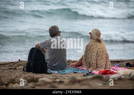 older or middle-aged couple sitting together on a beach on a stormy day with shawl wrapped around shoulders to protect from the wind, Stock Photo