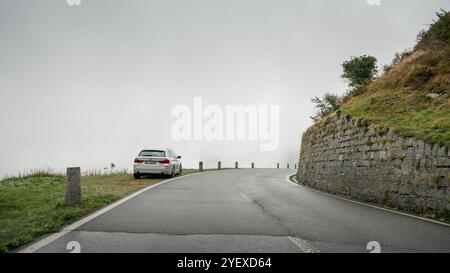 White BMW 5-series parked on a side of mountain pass in Swiss alps. Rear view of german car parked on a shoulder, road turning right, low visibility. Stock Photo