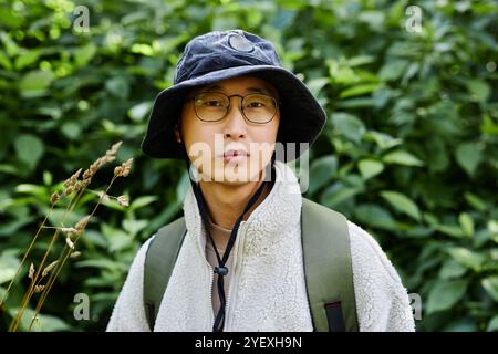 Vibrant portrait of young Asian man as male tourist looking at camera against forest greenery and wearing eyeglasses copy space Stock Photo