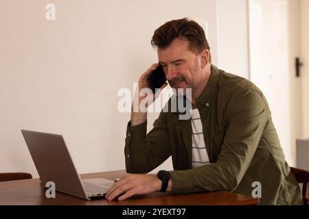 senior man using smartphone and laptop at home, smiling while working Stock Photo