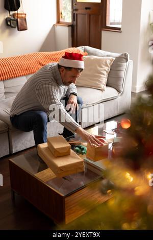 senior man in Santa hat wrapping Christmas gifts at home, feeling festive Stock Photo
