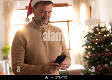 senior man in Santa hat using smartphone near Christmas tree at home Stock Photo