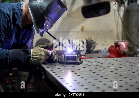 Metalworker wearing protective mask and gloves welding a custom mechanical part using a mig welder in a workshop Stock Photo