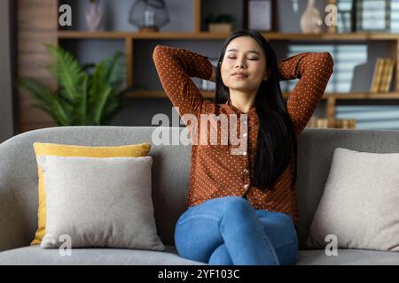 Asian woman relaxing at home on sofa, eyes closed, hands behind head. Embraces calm and peace, enjoying quiet moments in cozy living room. Concept of relaxation, tranquility. Stock Photo