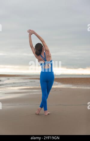 Rear view of a sporty woman standing doing an arm stretch exercise at the beach, preperation before swimming in the sea. Stock Photo