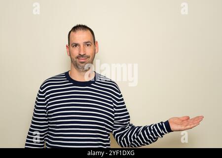 A bearded Hispanic man in his 40s wearing a striped sweater extending his hand to the side in a gesture of offering or outstretching, holding it palm Stock Photo