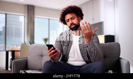 Worried Young Man Looking At Smartphone And Sitting In Front Of Sofa At Home Stock Photo