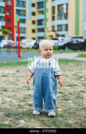 A 1-year-old baby boy in denim overalls is walking on the street. Children's oversized overalls. Stylish little boy on the playground. Stock Photo