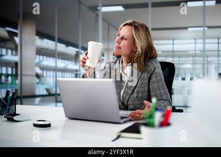 Businesswoman Working On Laptop At Office Desk, Drinking Coffee And Thinking Stock Photo
