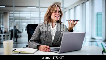 Professional Businesswoman Using Smartphone App For Client Call In Modern Office Setting With Laptop And Smiling Stock Photo