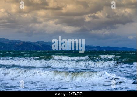 Waves roll relentlessly onto the sandy beach as clouds gather over distant mountains during a vibrant sunset. Stock Photo