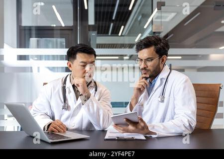 Two doctors engage in medical discussion, using laptop and tablet, in office. Concept of technology, collaboration, consultation. Stethoscope, healthcare professionals, teamwork, medical discussion. Stock Photo