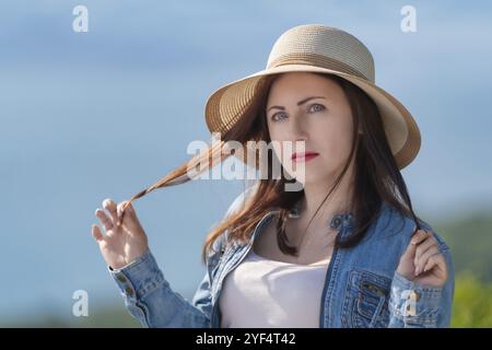 Mysterious adult woman with gray eyes looking at camera. Hipster dressed in straw hat, denim jacket and light T-shirt. Female tourist standing on blur Stock Photo