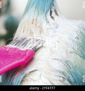 Close-up of stylist using pink brush while applying paint to female customer with emerald hair color, during process of bleaching hair roots in hair s Stock Photo