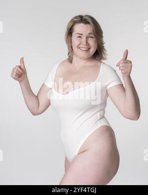 Portrait of plus size woman in bodysuit raising both hands and showing thumbs up, posing on white background. Caucasian extended sizes model is smilin Stock Photo
