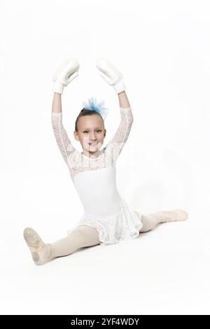 Smiling girl ballet dancer nine-year-old in white ballet tutu skirt, blue bow in hair and white mittens ballet stretching on white floor. Studio shot. Stock Photo