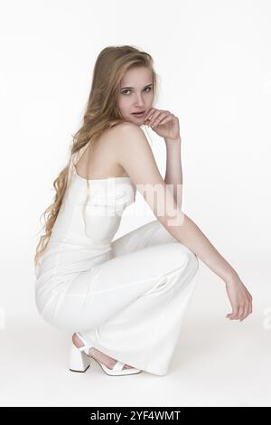 Blonde woman in white jumpsuit sitting squatting position raised hand to chin, put other on knee and looking over shoulder at camera. Side view of mod Stock Photo