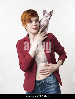Beautiful hipster redhead young woman looking at camera, dressed in red jacket, blue jeans and playful Sphynx Hairless kitten sitting on her shoulder. Stock Photo