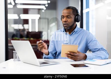 Mature african american businessman in video conference, wearing headphones, holding notepad while talking to laptop. Engaged in virtual meeting in modern office. Represents remote work, technology Stock Photo