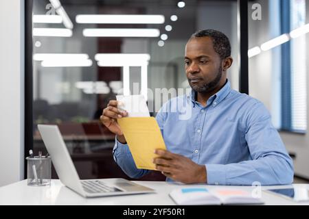 Mature African American businessman in office using laptop, opening envelope. Focused expression suggests concentration, professionalism. Bright lighting, modern setting denotes corporate environment Stock Photo