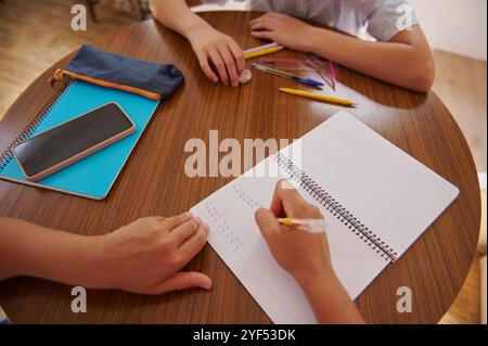 Two students collaborate on a study project at a round wooden table, with notebooks, pens, and a smartphone scattered around. They focus on learning a Stock Photo