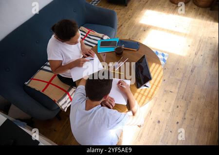 Overhead view of two young individuals collaborating on writing tasks at a wooden table in a cozy room, surrounded by gadgets and stationery. Stock Photo