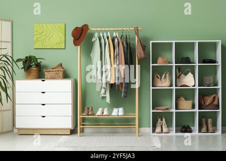 Stylish autumn clothes hanging on rack, chest of drawers and shelving unit against green wall Stock Photo