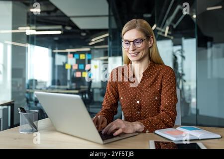 Mature business woman working on laptop in modern office setting. She appears professional and focused, exuding confidence amidst workspace with notes. Ideal scene for business success and motivation Stock Photo