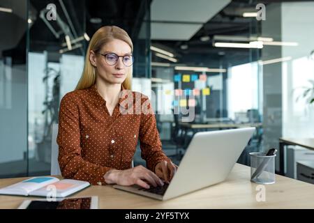 Serious businesswoman working on laptop at office. Mature professional focused on important tasks. Business attire and organized workspace highlight dedication and expertise in modern environment. Stock Photo
