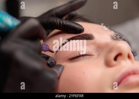 Beautician in black gloves applying permanent makeup on eyebrows using a tattoo machine Stock Photo