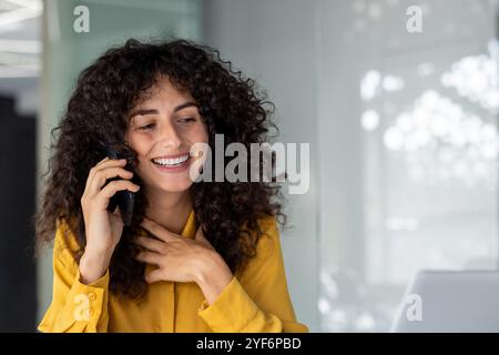 Smiling woman in yellow shirt talking on smartphone, conveying joyful conversation and connection. Modern office setting enhances communication theme. Bright, positive ambiance with friendly vibe. Stock Photo