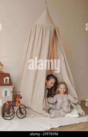 Happy mother and daughter sitting in a play teepee next to a dolls house in a bedroom Stock Photo