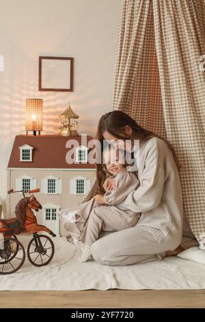 Happy mother and daughter sitting in a play teepee next to a doll's house in a bedroom Stock Photo