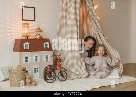 Happy mother and daughter sitting in a play teepee next to a doll's house in a bedroom Stock Photo